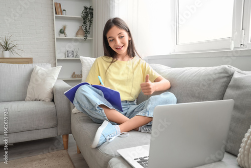 Little schoolgirl with copybook and laptop studying online on sofa at home