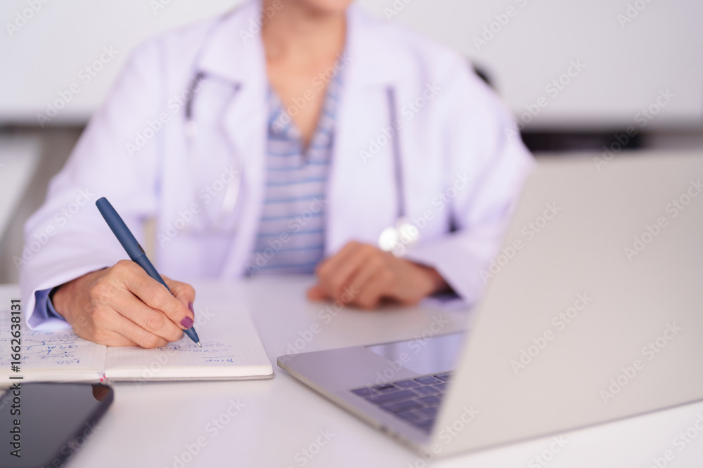 Fototapeta premium Experienced female doctor writes patient notes medical records consultation details on office desk with laptop smartphone healthcare provider at clinic