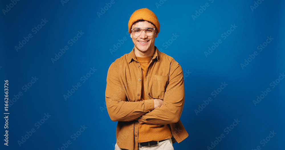 Obraz premium Confidence, portrait and man in studio for fashion, smile and happiness on mockup space. Arms crossed, style and male model with trendy clothes, glasses and gen z outfit on blue background in Spain