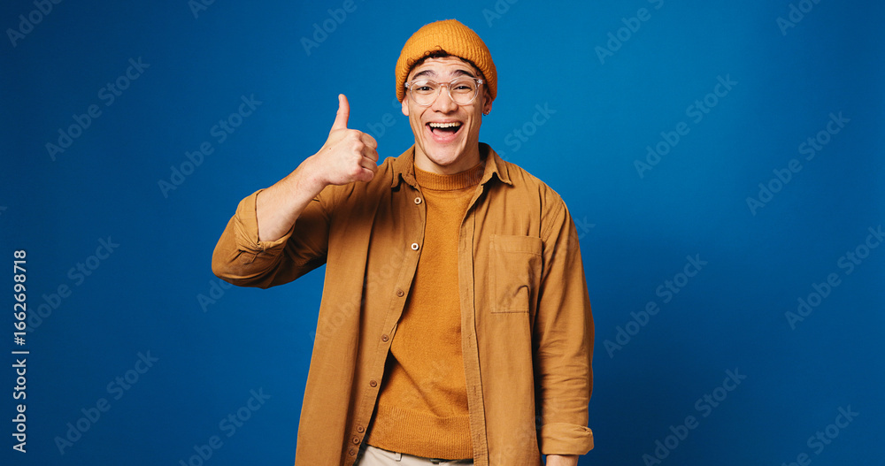 Naklejka premium Happy man, portrait and fashion with thumbs up in studio for good choice on a blue background. Male person, model or gen z with smile, like emoji or yes sign for style or clothing on mockup space