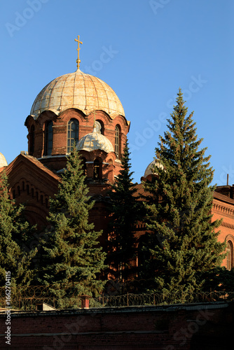 Wallpaper Mural Historic red-brick prison church “Kresty” in Saint Petersburg with golden-brown domes, golden cross, evergreen trees, and barbed wire under sunny sky. Torontodigital.ca