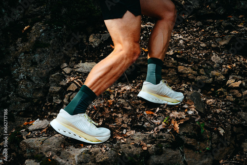 Brand new white trail running shoes on rocky uphill terrain with a professional runner on rugged natural ground, close-up on muscular legs