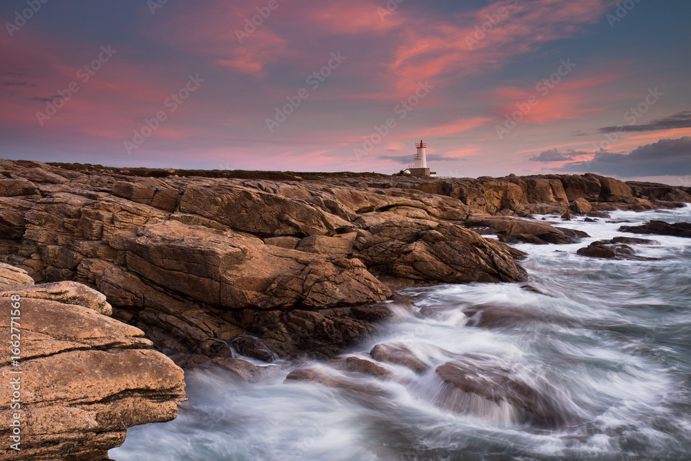Obraz premium Lorient Morbihan Waves rushing against the rocks with a lighthouse in the background during sunset.