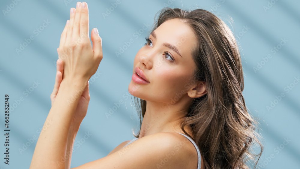 Fototapeta premium Beautiful young woman with healthy skin gently applying skincare product to her hands, showcasing a radiant complexion against a soft blue background, emphasizing the importance of skincare 