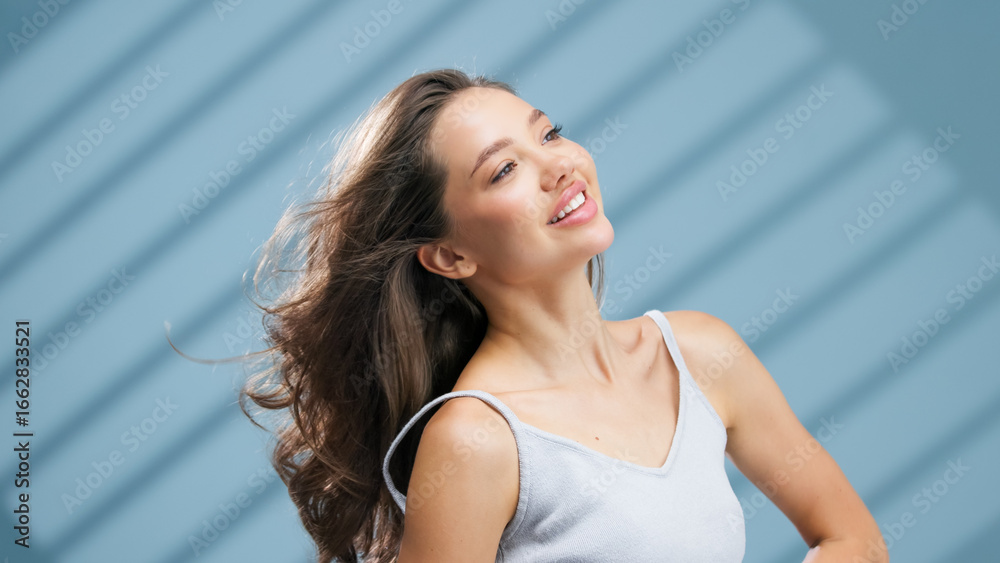 Fototapeta premium Young woman with healthy skin and flowing hair, smiling joyfully against a soft blue background with light shadows, embodying the essence of skincare and beauty in a vibrant atmosphere