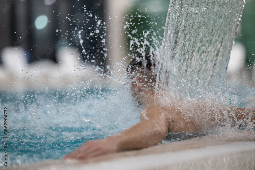 Fototapeta premium Man standing under a hydrotherapy waterfall in a spa pool, receiving water massage on his head and shoulders.