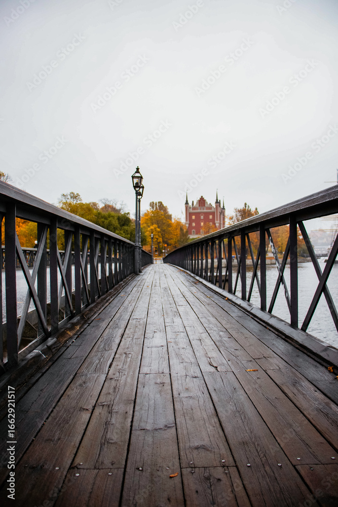 Obraz premium A wooden bridge leading across a river to a historic red building on the opposite bank, surrounded by colorful autumn trees in Stockholm, Sweden.