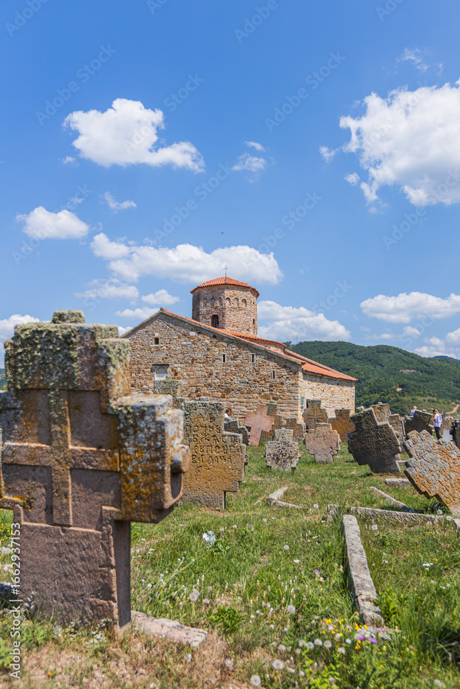 Fototapeta premium The ancient stone Church of St. Peter, a prominent Serbian Orthodox landmark, is framed by medieval tombstones and a clear blue sky with white clouds, historical and spiritual landscape in Novi Pazar.
