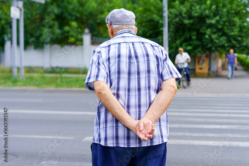 Elderly man waits to cross a busy street with hands clasped behind his back during daylight