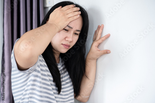 Dizzy Asian woman standing at home, leaning on the wall for support, suffering from vertigo, headache, and migraine pain, feeling spinning, unsteady, and weak.