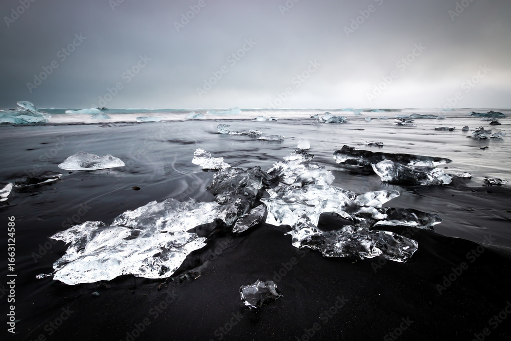 Obraz premium Long exposure of crystal-clear chunks of ice lying on Diamond Beach black sand of Iceland’s coast near Jökulsárlón, creating a striking contrast in color and texture