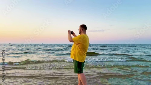Young man filming the sea with his smartphone. Calm seaside moment capturing nature and waves on a sunny day
