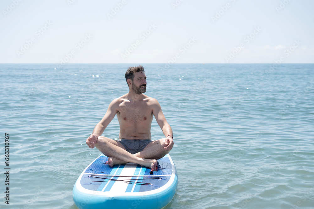 Fototapeta premium Man meditating on paddle board in ocean practicing yoga