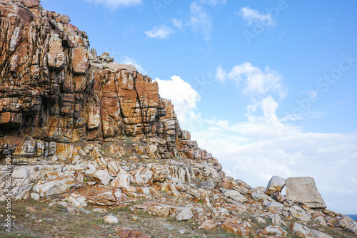 A striking view of rugged rocky cliffs with layered textures and vibrant earthy tones, set against a bright blue sky with scattered clouds. Captured at the travel, and adventure at Cape of Good Hope
