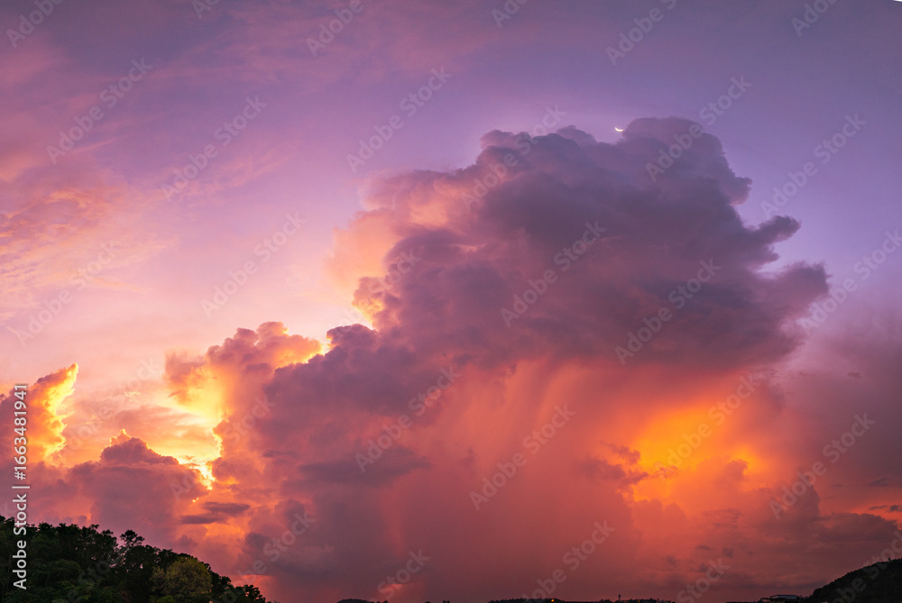 Fototapeta premium A vivid and dynamic sky captures towering storm clouds illuminated by the warm hues of a setting sun, creating a striking contrast between light and shadow above a darkened forested landscape.