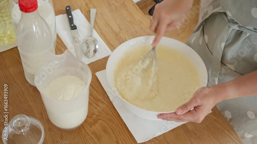 A woman intensively whisks the batter in a white bowl with a metal whisk to remove all the lumps. She prepares a smooth, uniform dough for cooking delicious homemade pancakes or crepes.