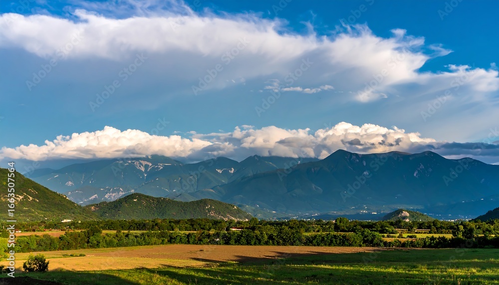 Obraz premium Panoramic view of a valley with rolling hills and mountains under a vibrant blue sky dotted with fluffy white clouds.