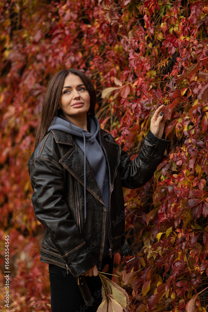 Beautiful young brunette girl walks in autumn park against the background of a wall with red leaves. autumn concept. autumn season. fall