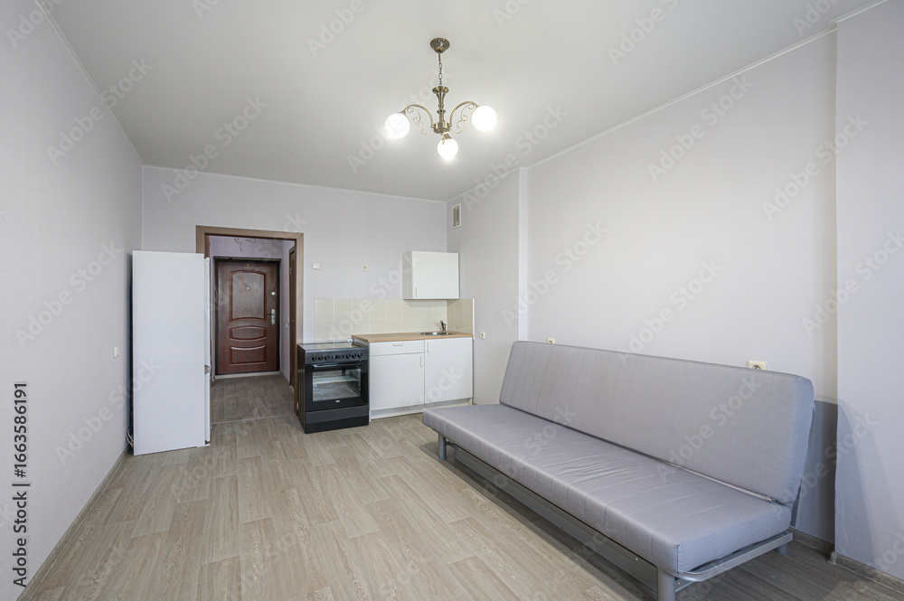 Fototapeta premium simple studio apartment. Features a sofa, kitchenette with stove, and a fridge. Walls are light gray, floor is laminate