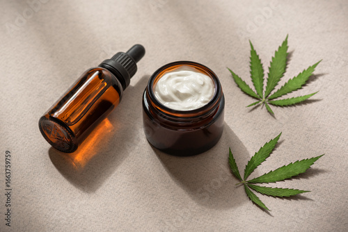 Face cream with cannabis and oil serum in an amber dropper bottle. THC tincture, CBD oil and hemp leaves on a beige paper background