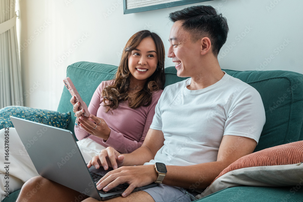 Fototapeta premium Young Asian couple sitting on green sofa at home, smiling while using laptop and smartphone together. Woman wears pink top, man in white t-shirt with smartwatch, cozy indoor background