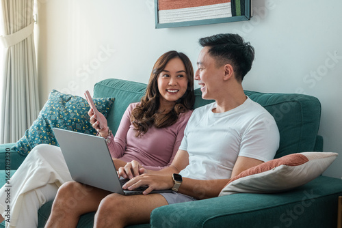 Young Asian couple sitting on green sofa at home, smiling while using laptop and smartphone together. Woman wears pink top, man in white t-shirt with smartwatch, cozy indoor background