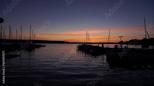 Moored boats in the marina of Berlin’s Lake Wannsee at dusk, reflecting the warm tones of a vivid orange and purple sunset.