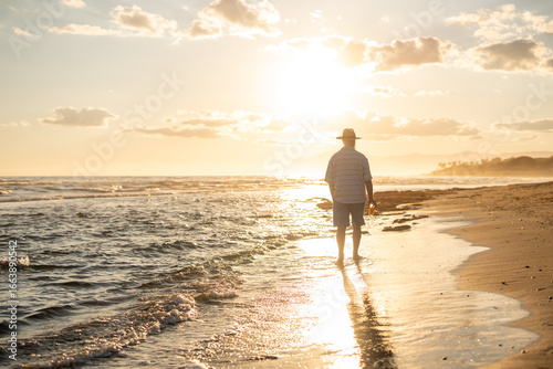 Canvas Print Elderly man in a straw hat walking barefoot along the beach shoreline at golden hour, holding a glass of orange juice and enjoying the peaceful sunset over the calm sea