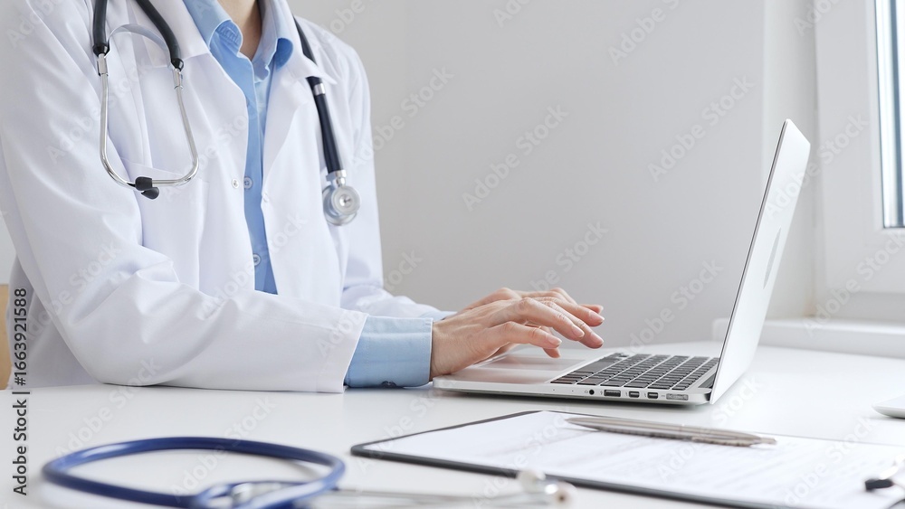 Doctor woman is typing on a laptop with a stethoscope, medical record, and pen on the desk in a bright medical office, showcasing healthcare technology and medical documentation. Medicine concept