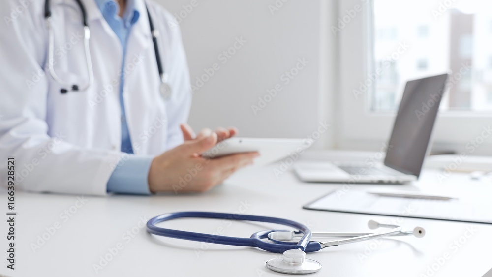 Closeup of blue stethoscope is lying on the table near female doctor reviewing prescription on digital tablet, representing digital healthcare workflow in contemporary medical setting. Medicine