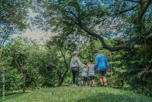 A beautiful moment of parenting. An Asian mother and father guide their two young sons by the hand up a green hill, creating cherished childhood memories in a beautiful park setting.