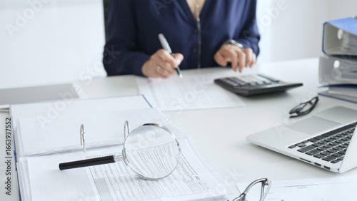 Tableau sur toile Close up of magnifying glass and calculator over financial documents opposite female accountant analyzing data, reviewing reports, binders and laptop on a white desk