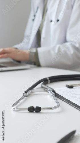 Stethoscope and medical records are on the desk opposite the doctor is typing on a laptop with a in a medical office, suggesting online healthcare or telemedicine services. Medicine concept