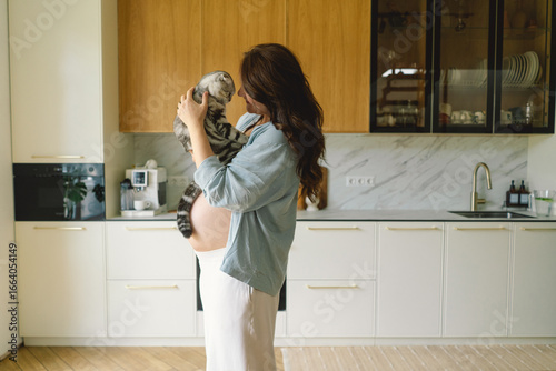 In a contemporary kitchen, a pregnant woman smiles while holding her cat close to her baby bump. Warm wooden cabinets create a cozy environment for this heartwarming moment.