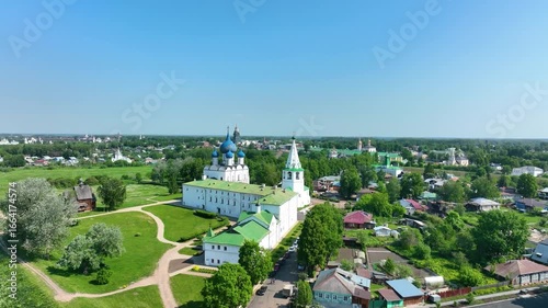 A beautiful, wide panoramic view of a famous historic kremlin, including a cathedral with blue domes, set in a green park overlooking a provincial town on a sunny day