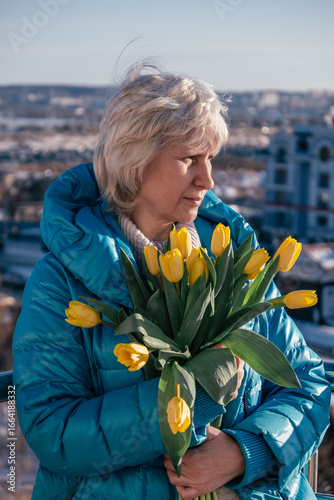 authentic portrait woman with short blonde hair stands outdoors in blue jacket, holding bright yellow tulips. cityscape is visible in background with clear and sunny day. vertical. close up.