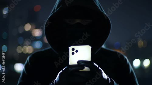 Anonymous hooded hacker in a mask using a smartphone at night with city lights background
