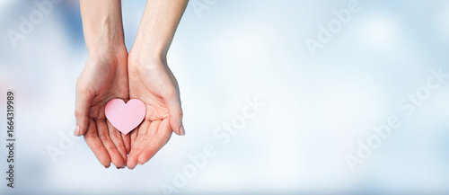 Obraz na płótnie A pair of human hands gently holding a pink heart shape against a soft blue background