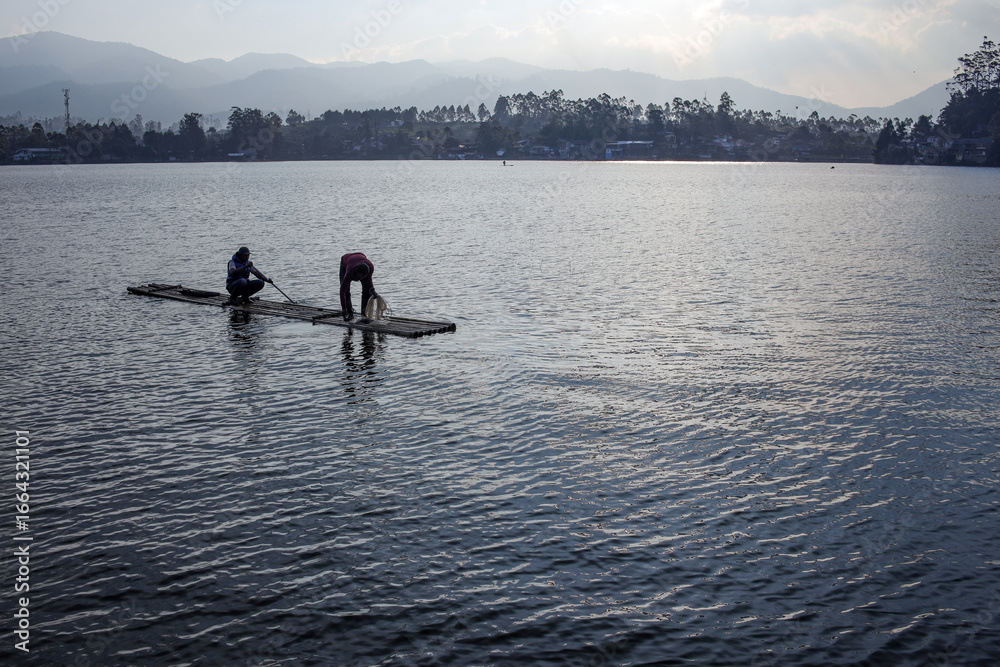 Fototapeta premium Two Fishermen On A Bamboo Raft, Preparing Their Nets On The Lake, With Mountains In The Background
