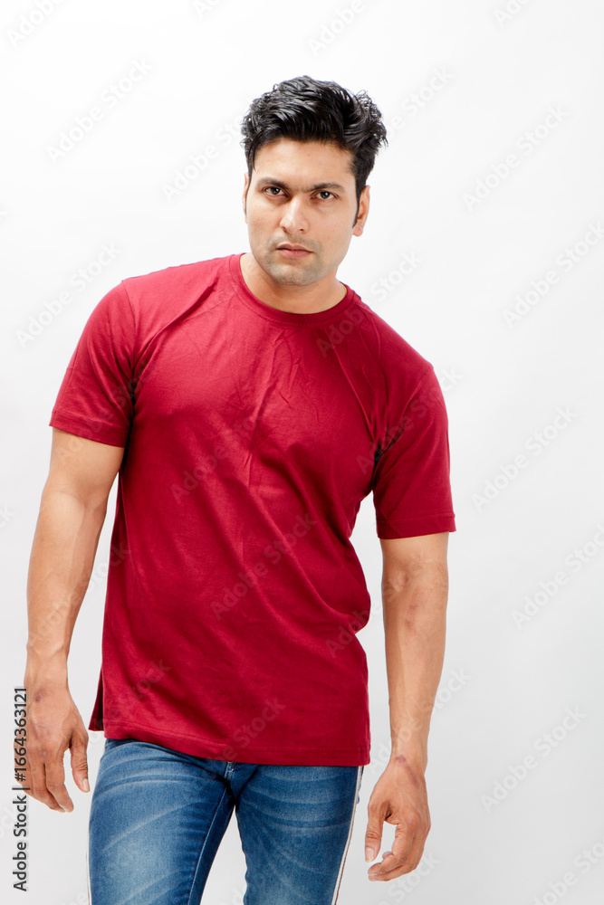 Fototapeta premium Young man wearing a red t-shirt, denim jeans, and sneakers posing casually in a studio against a plain white background. Ideal for themes of fashion, confidence, and modern lifestyle.