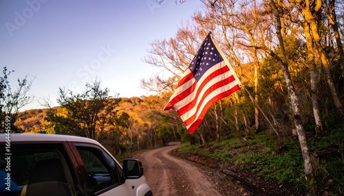 A vibrant American flag waves gently over a dusty forest road, highlighting the golden hues of the late afternoon sunlight.
