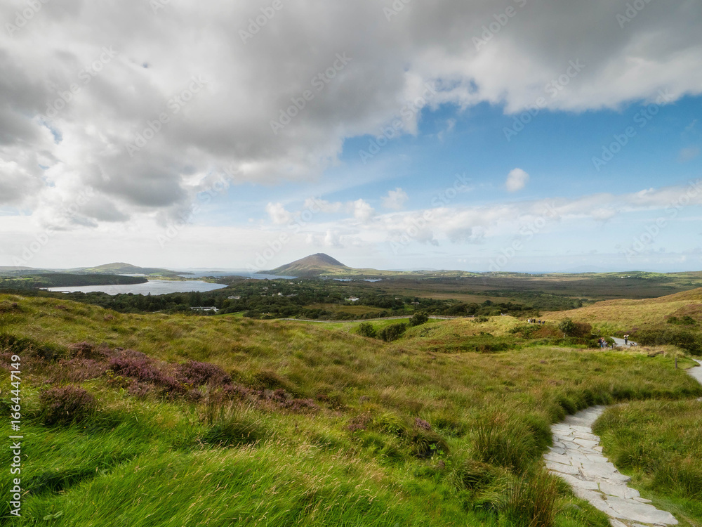 Fototapeta premium A lush green field with a mountain and ocean in the background. The sky is clear and blue. Beautiful nature view from Diamond hill, Connemara National Park, Ireland. Travel and tourism.