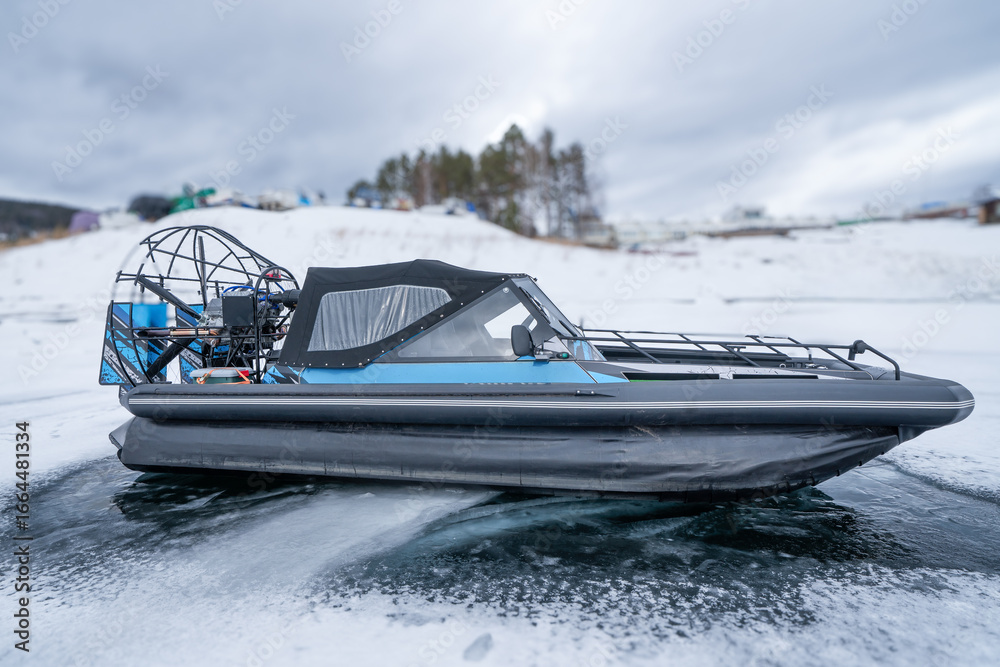 Fototapeta premium A modern hovercraft glides effortlessly over a frozen lake, showcasing its sleek design. The surrounding landscape is a serene winter scene, with snow-covered hills in the background and scattered