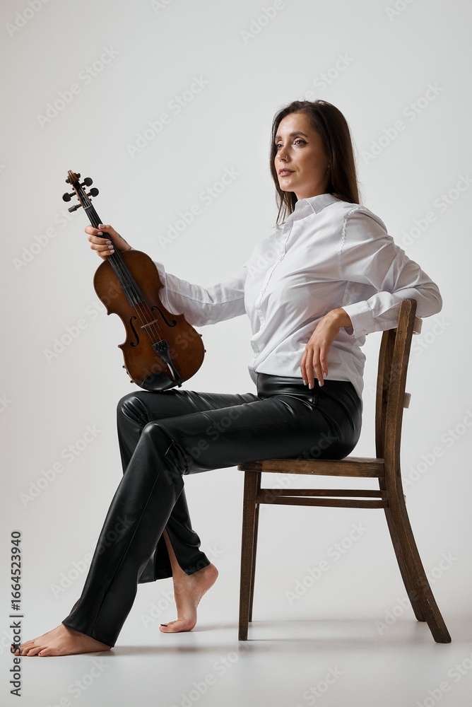 Fototapeta premium A woman with a violin in a white shirt and black pants is seated in a wooden chair.