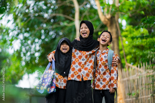 Three indonesia children happily walking together on a village road, wearing matching batik clothes and carrying school bags. Joyful friendship and family moment.