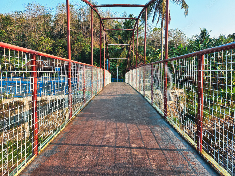 Obraz premium A striking red metal pedestrian bridge with a worn walkway and wire mesh fence leading through a lush green tropical forest on a bright sunny day
