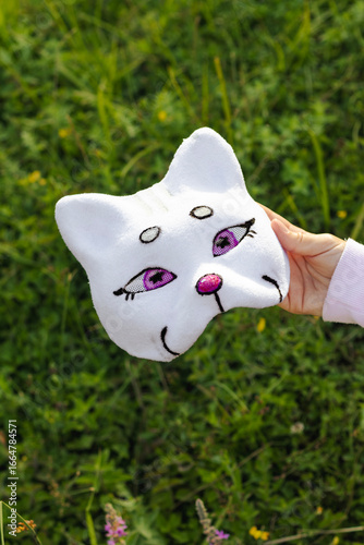 Close-up of white plush cat mask with purple eyes and pink nose held in hand over grass and flowers. Quadrobics subculture, youth hobby, animal mimicry, playful identity accessory.
