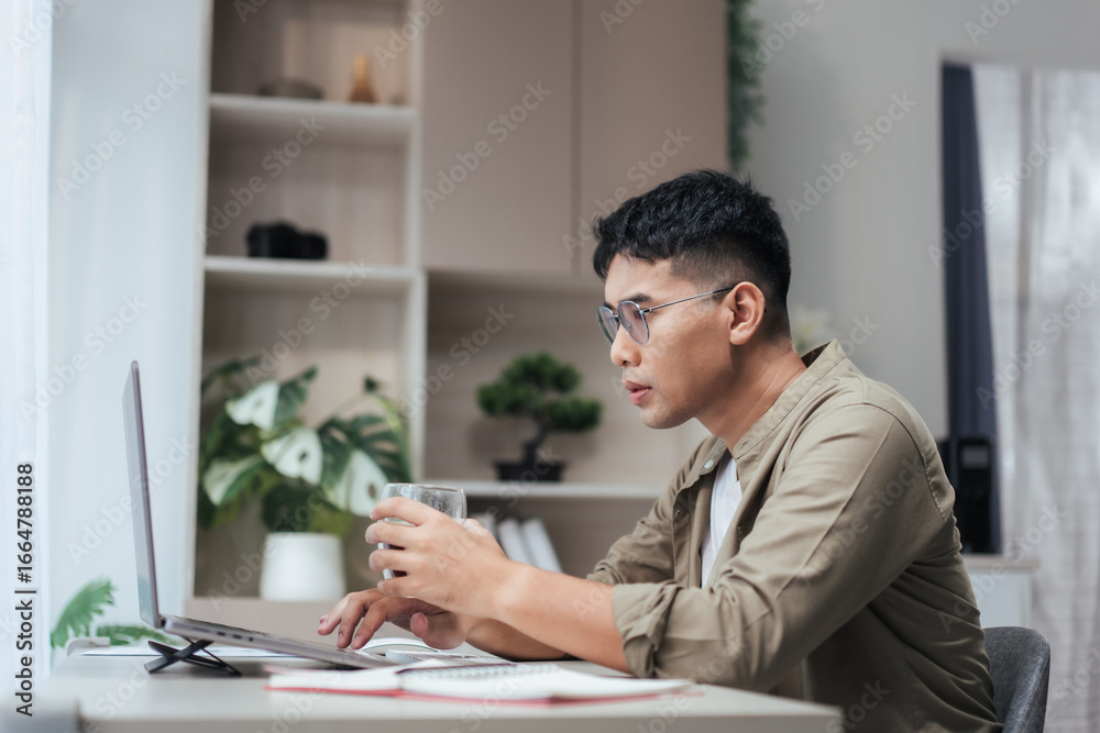 Fototapeta premium Asian man at a home office looks at his laptop while holding a coffee glass. Open notebook and pen on the desk, with soft daylight and green plants creating a neat, modern space for remote work.