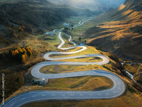 Aerial view of winding mountain road, river in alpine valley at sunset in autumn. Top drone view of road in Swiss mountain pass, yellow grass, trees in fall. Colorful landscape. Alps in Switzerland