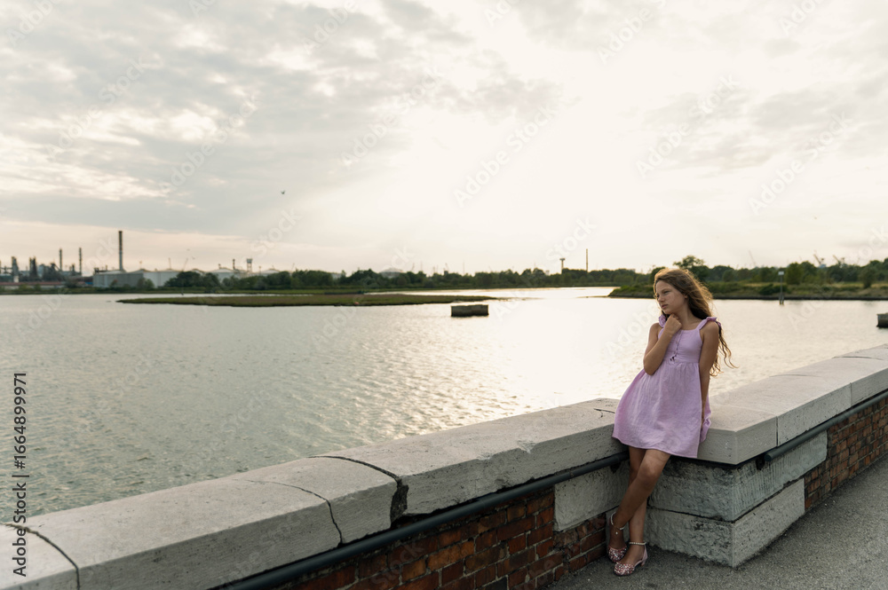 Full body of a young girl in a purple dress resting on the side of Ponte della Libertà in Venice, Italy, looking toward the lagoon.